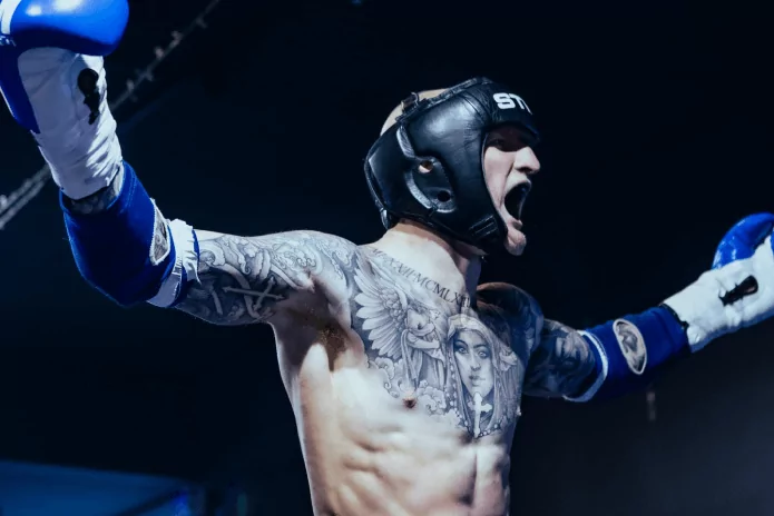 Tattooed martial artist in headgear raises arms in victory with blue gloves