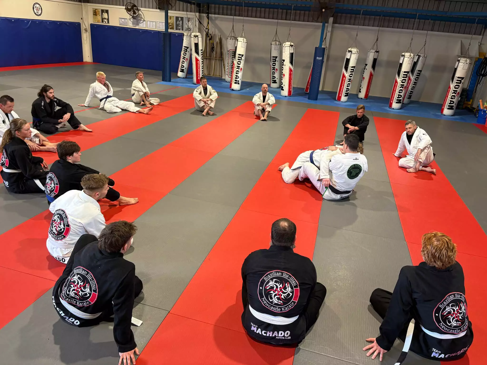 Students seated in a circle attentively watch BJJ grappling technique demonstration on mats